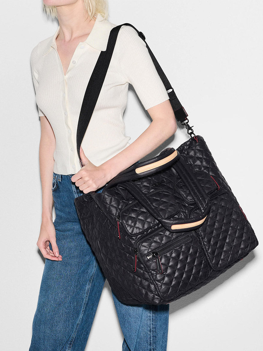 A person wearing a short-sleeve white shirt and blue jeans carries the MZ Wallace Large Metro Travel Tote in Black Oxford.