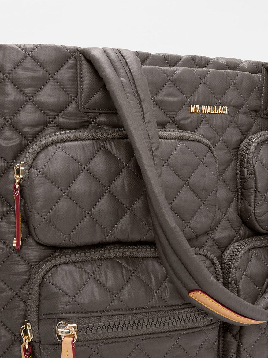 Close-up of the MZ Wallace Large Metro Travel Tote in Magnet Oxford, a quilted dark gray bag with durable Oxford fabric, gold "MZ WALLACE" branding, and zippered pockets with red and tan pulls.