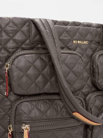Close-up of the MZ Wallace Large Metro Travel Tote in Magnet Oxford, a quilted dark gray bag with durable Oxford fabric, gold "MZ WALLACE" branding, and zippered pockets with red and tan pulls.