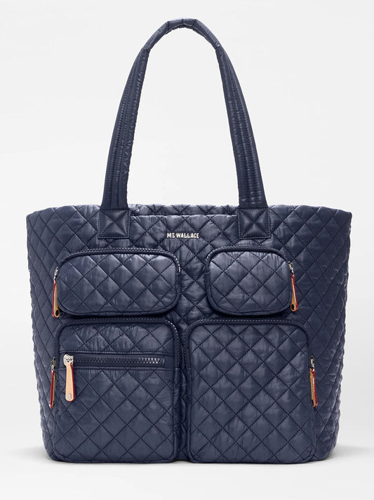 The MZ Wallace Large Metro Travel Tote in Dawn Oxford is a navy blue quilted bag made from recycled nylon, with double handles and four front zippered pockets accented by bright orange zipper pulls.