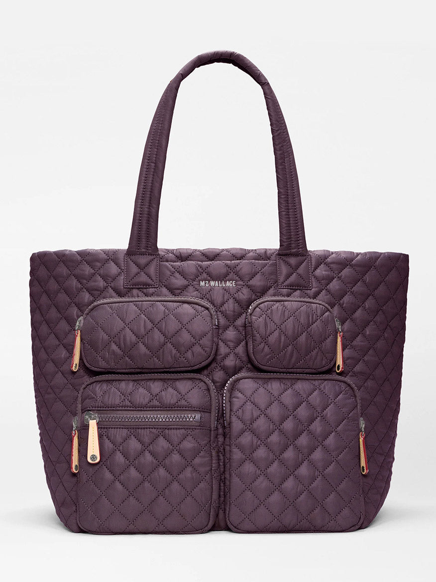 The MZ Wallace Large Metro Travel Tote in Raisin Oxford is a quilted purple bag made from recycled nylon, designed with four zippered exterior pockets for organized storage and two sturdy handles, and features subtle “MZ WALLACE” branding near the top center.