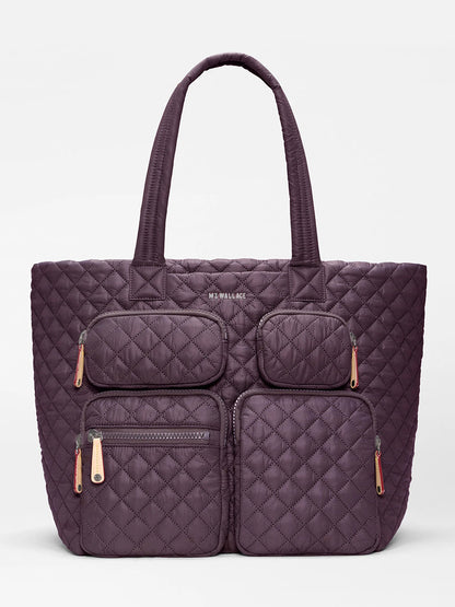 The MZ Wallace Large Metro Travel Tote in Raisin Oxford is a quilted purple bag made from recycled nylon, designed with four zippered exterior pockets for organized storage and two sturdy handles, and features subtle “MZ WALLACE” branding near the top center.