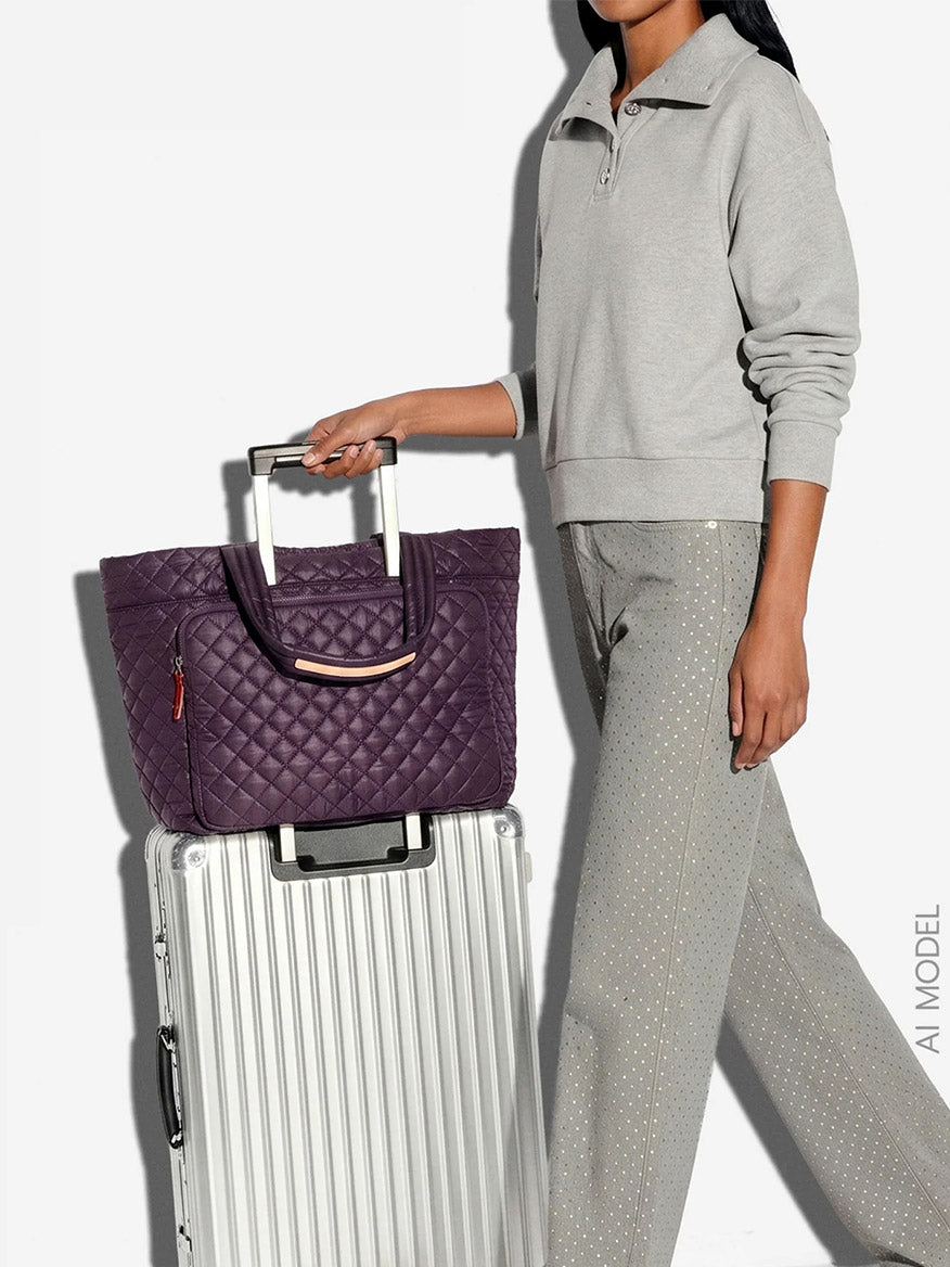 Dressed in light gray, a person stands with the MZ Wallace Large Metro Travel Tote in Raisin Oxford—crafted from recycled nylon—in a quilted purple design atop a silver rolling suitcase, presenting an organized carry against a plain white background.