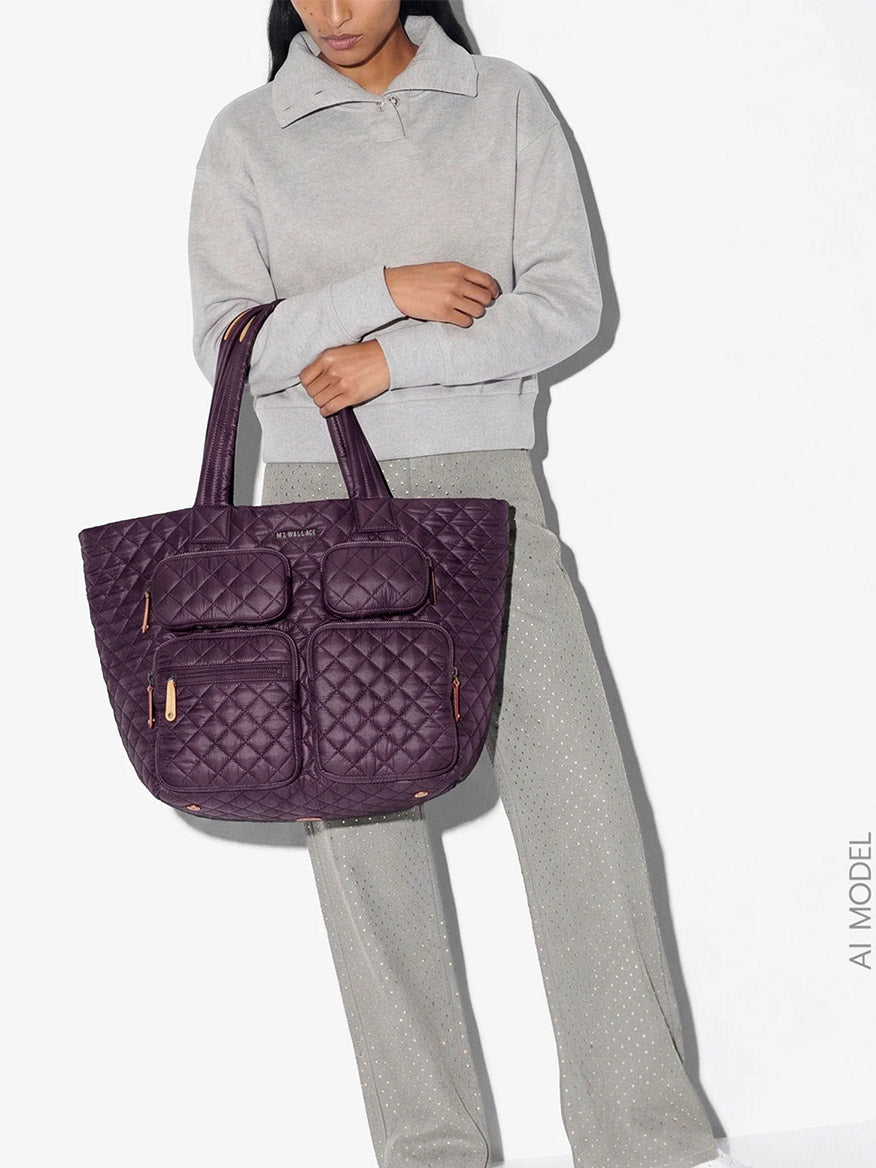 A person wearing a light gray sweater and gray pants holds the MZ Wallace Large Metro Travel Tote in Raisin Oxford, a spacious quilted bag featuring multiple external pockets for organized carry. Crafted from recycled nylon, this travel tote delivers eco-friendly style.