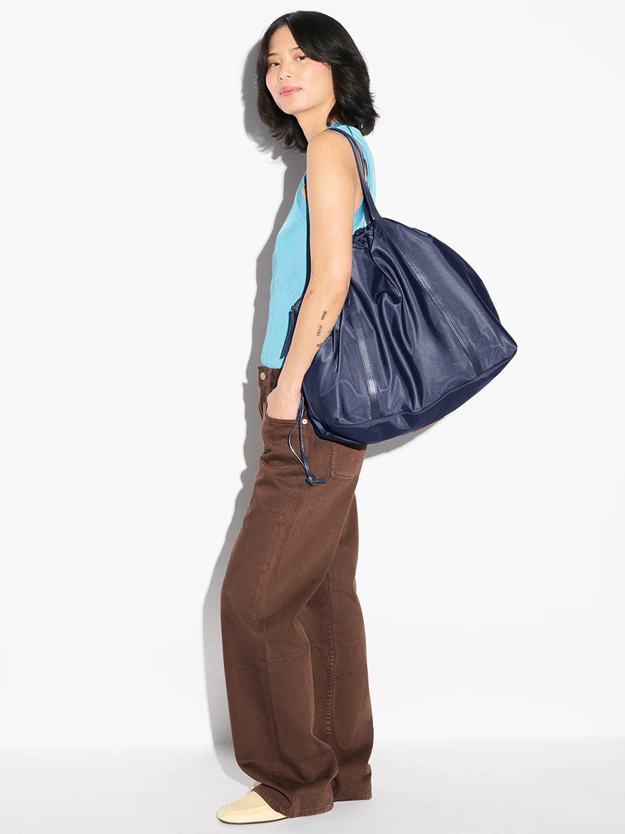 A woman in a blue sleeveless top, brown pants, and cream shoes stands sideways carrying the MZ Wallace Large Waverly Tote in Dawn Bedford with a crossbody strap.