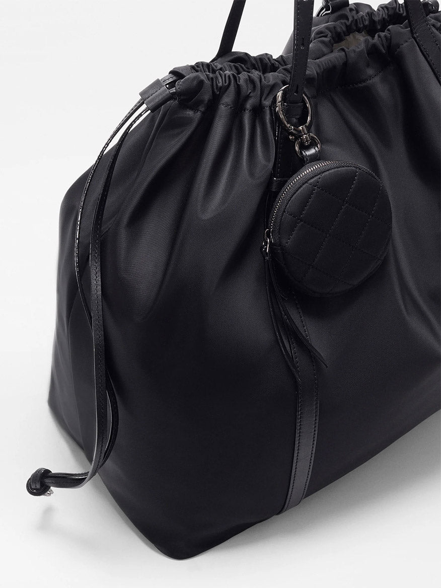 The MZ Wallace Large Waverly Tote in Black Bedford is a spacious black drawstring bag with Italian leather handles, a round quilted coin pouch on the handle, fits a 15" laptop, and is shown on a plain white background.