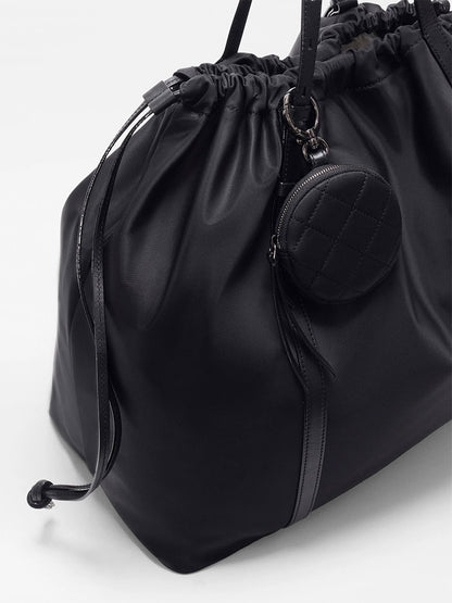 The MZ Wallace Large Waverly Tote in Black Bedford is a spacious black drawstring bag with Italian leather handles, a round quilted coin pouch on the handle, fits a 15" laptop, and is shown on a plain white background.