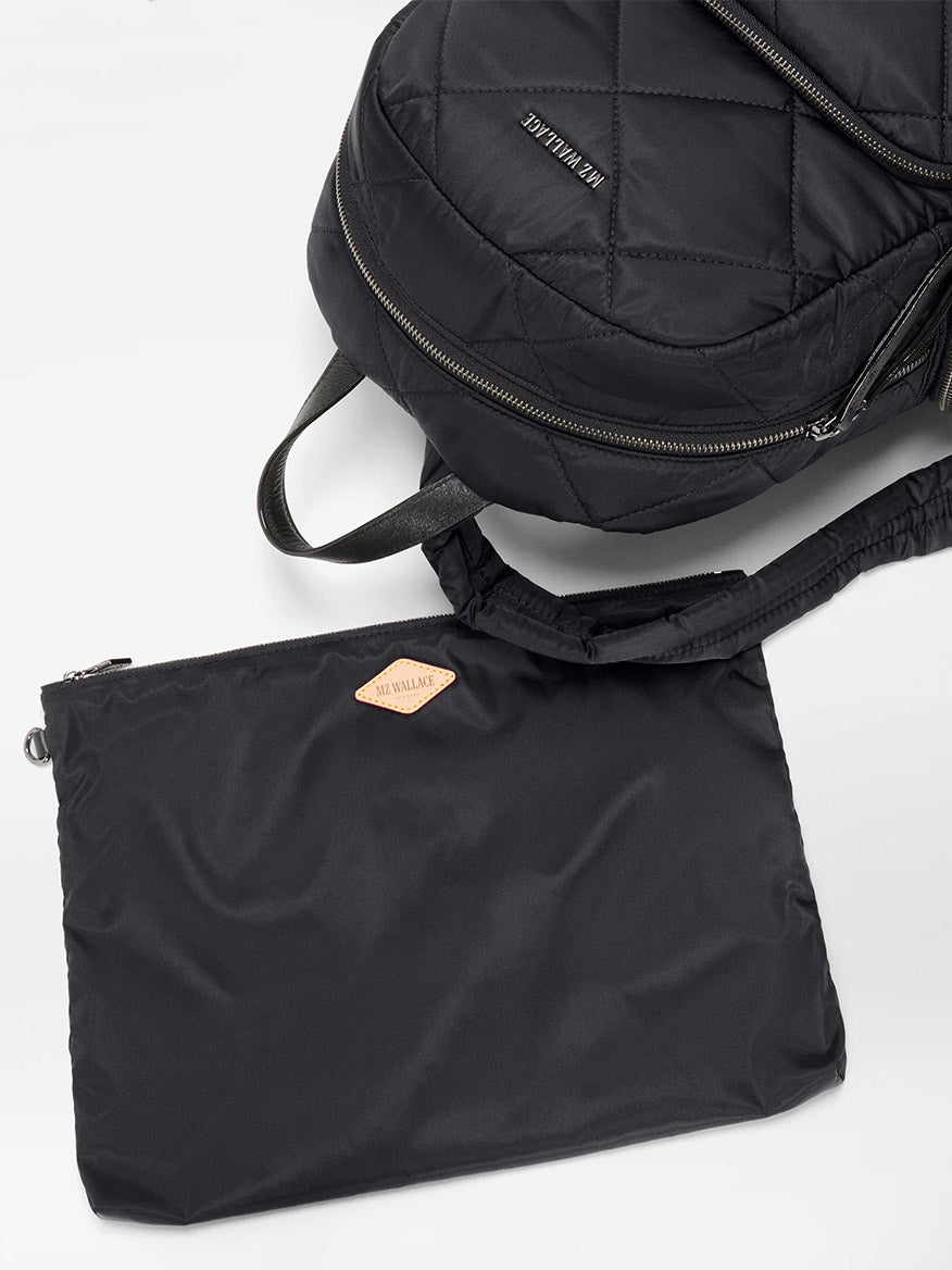 The MZ Wallace Madison Backpack in Black Bedford, featuring quilted nylon, adjustable straps, and silver zippers, sits beside a matching black zippered pouch on a white surface.