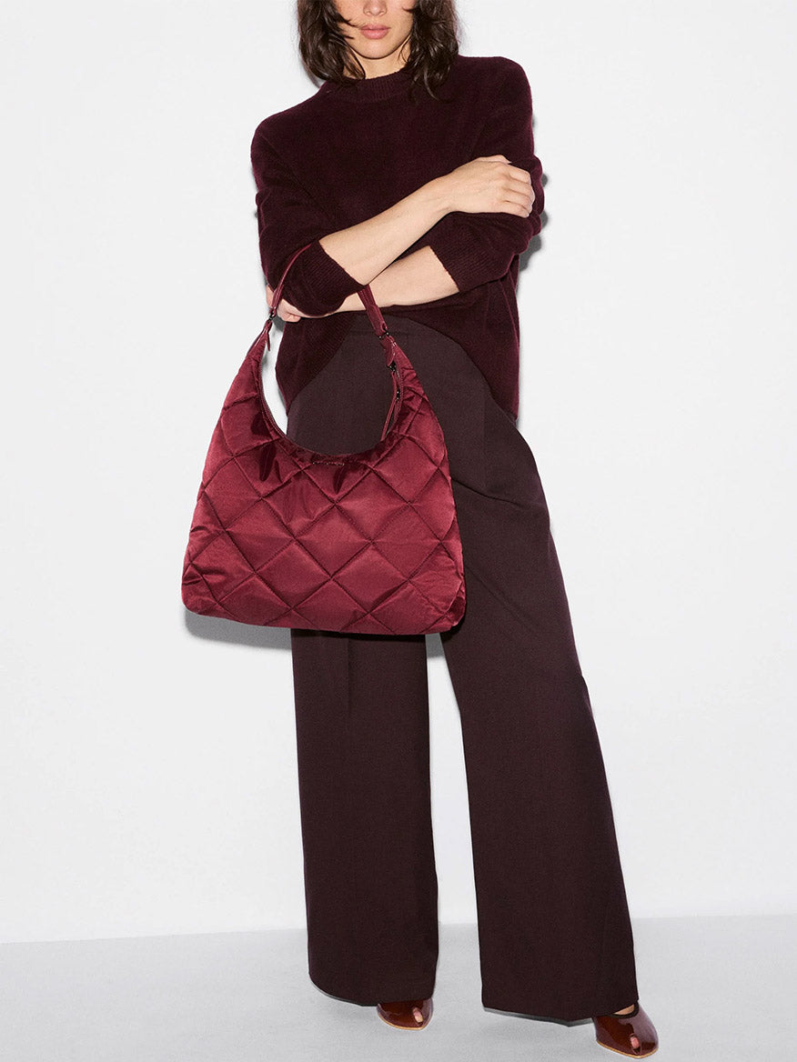 A woman stands against a white background wearing a dark red top and pants, holding the MZ Wallace Madison Shoulder in Claret Bedford—a large quilted burgundy Italian leather shoulder bag.