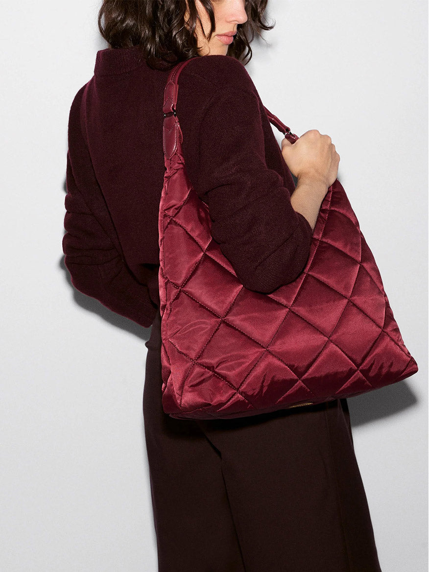 A woman, dressed in a dark burgundy top and trousers, stands sideways against a plain light background while holding the MZ Wallace Madison Shoulder in Claret Bedford—a large, quilted shoulder bag crafted from recycled nylon.