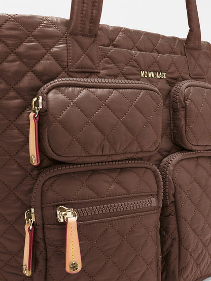 Close-up of the MZ Wallace Medium Metro Travel Tote in Walnut Oxford, a brown quilted bag with gold zippers and three front pockets, featuring “MZ WALLACE” branding in gold letters near the top.