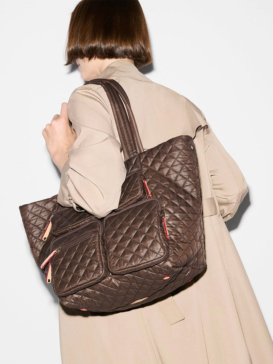 A person in a beige trench coat carries the MZ Wallace Medium Metro Travel Tote in Walnut Oxford, a large quilted brown recycled nylon bag with multiple zippered pockets, over their shoulder.