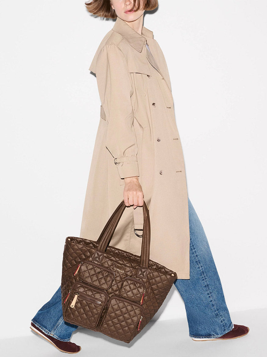 A person in a beige trench coat and blue jeans is holding the MZ Wallace Medium Metro Travel Tote in Walnut Oxford, featuring front pockets, while walking against a plain white background.