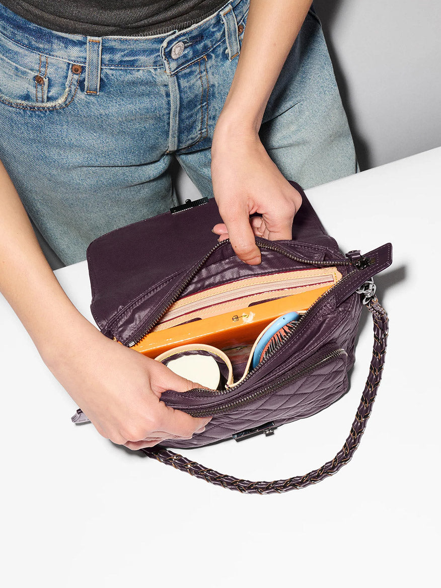 Wearing jeans and a gray shirt, a person opens the MZ Wallace Crosby Lock Medium Crossbody in Raisin Oxford—crafted from purple quilted recycled nylon with Italian leather trim—to reveal a compact, hairbrush, lip balm, and other small essentials on a white surface.