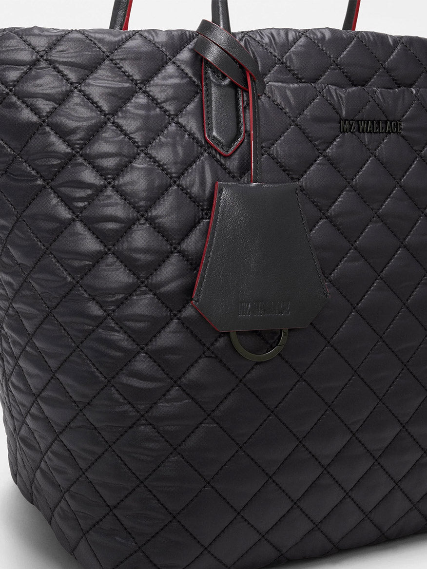 Close-up of the MZ Wallace Medium Daily Tote in Black Oxford, a black quilted tote with red trim and “MZ WALLACE” branding, featuring a leather tag on the handle. This versatile laptop bag is crafted from recycled nylon for a stylish and eco-friendly finish.