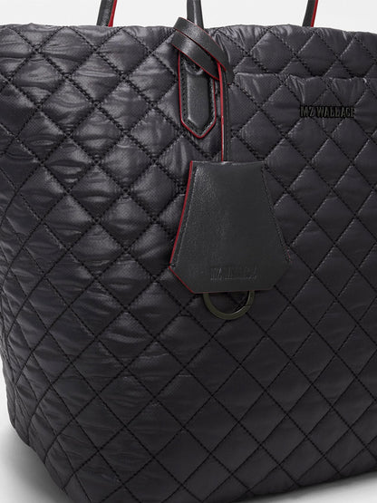 Close-up of the MZ Wallace Medium Daily Tote in Black Oxford, a black quilted tote with red trim and “MZ WALLACE” branding, featuring a leather tag on the handle. This versatile laptop bag is crafted from recycled nylon for a stylish and eco-friendly finish.