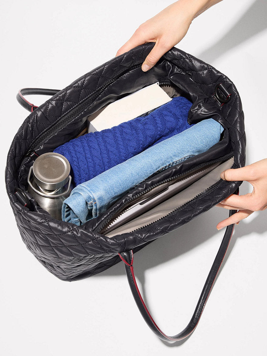 The MZ Wallace Medium Daily Tote in Black Oxford is shown open from above, containing a blue sweater, a book, a metal water bottle, rolled jeans, and a folder. This versatile laptop tote bag is perfect for carrying your daily essentials in style.