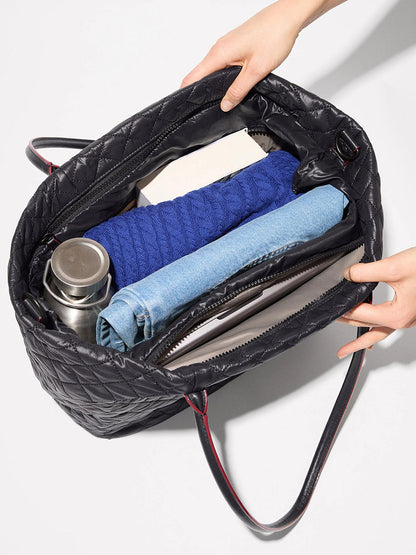The MZ Wallace Medium Daily Tote in Black Oxford is shown open from above, containing a blue sweater, a book, a metal water bottle, rolled jeans, and a folder. This versatile laptop tote bag is perfect for carrying your daily essentials in style.