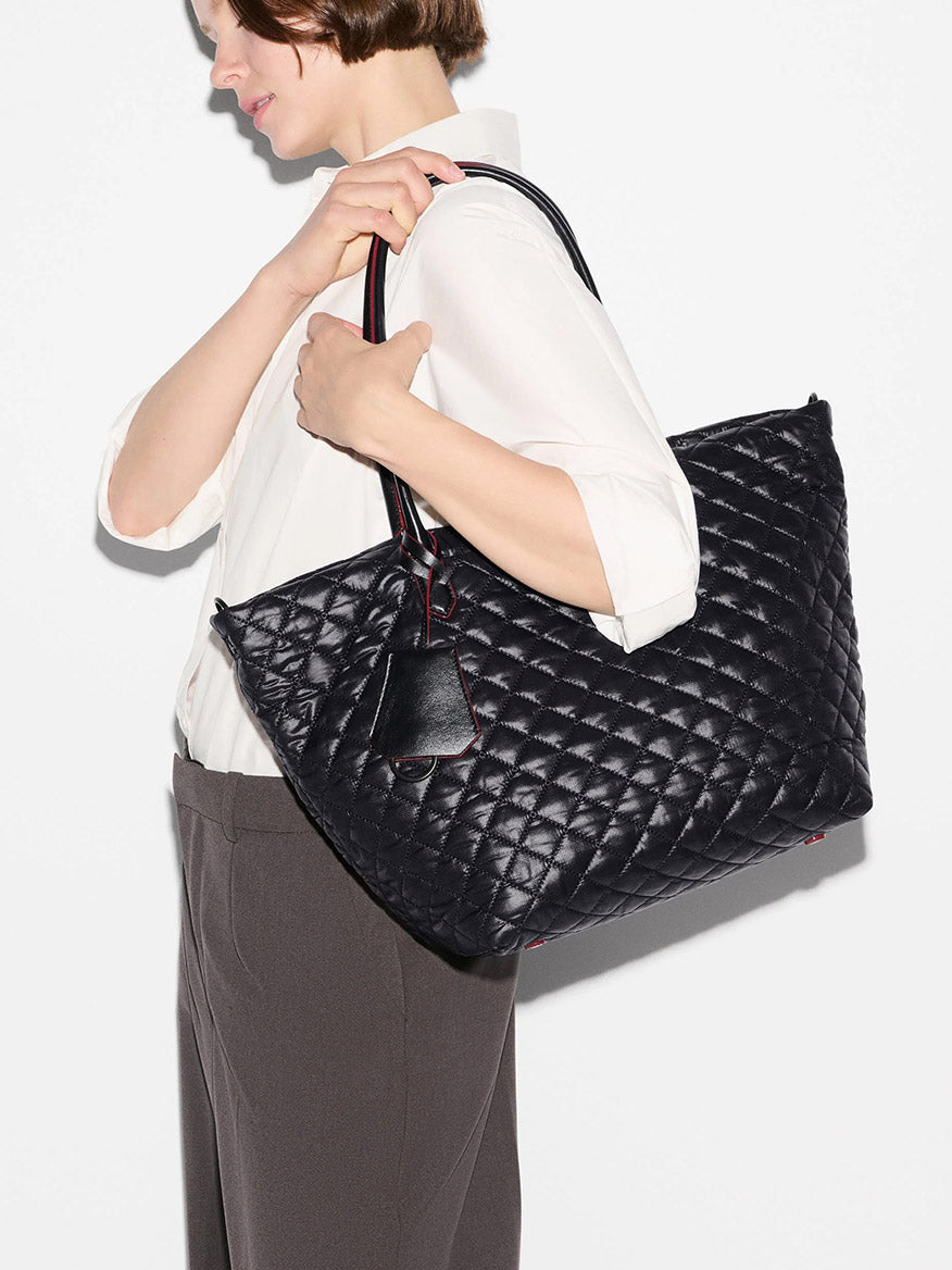 A person in a white shirt and dark pants holds the MZ Wallace Medium Daily Tote in Black Oxford over their shoulder against a plain background.