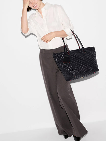 A person wearing a white shirt and wide-legged dark trousers holds the MZ Wallace Medium Daily Tote in Black Oxford—a sophisticated, spacious black quilted tote bag—while standing against a plain white background.
