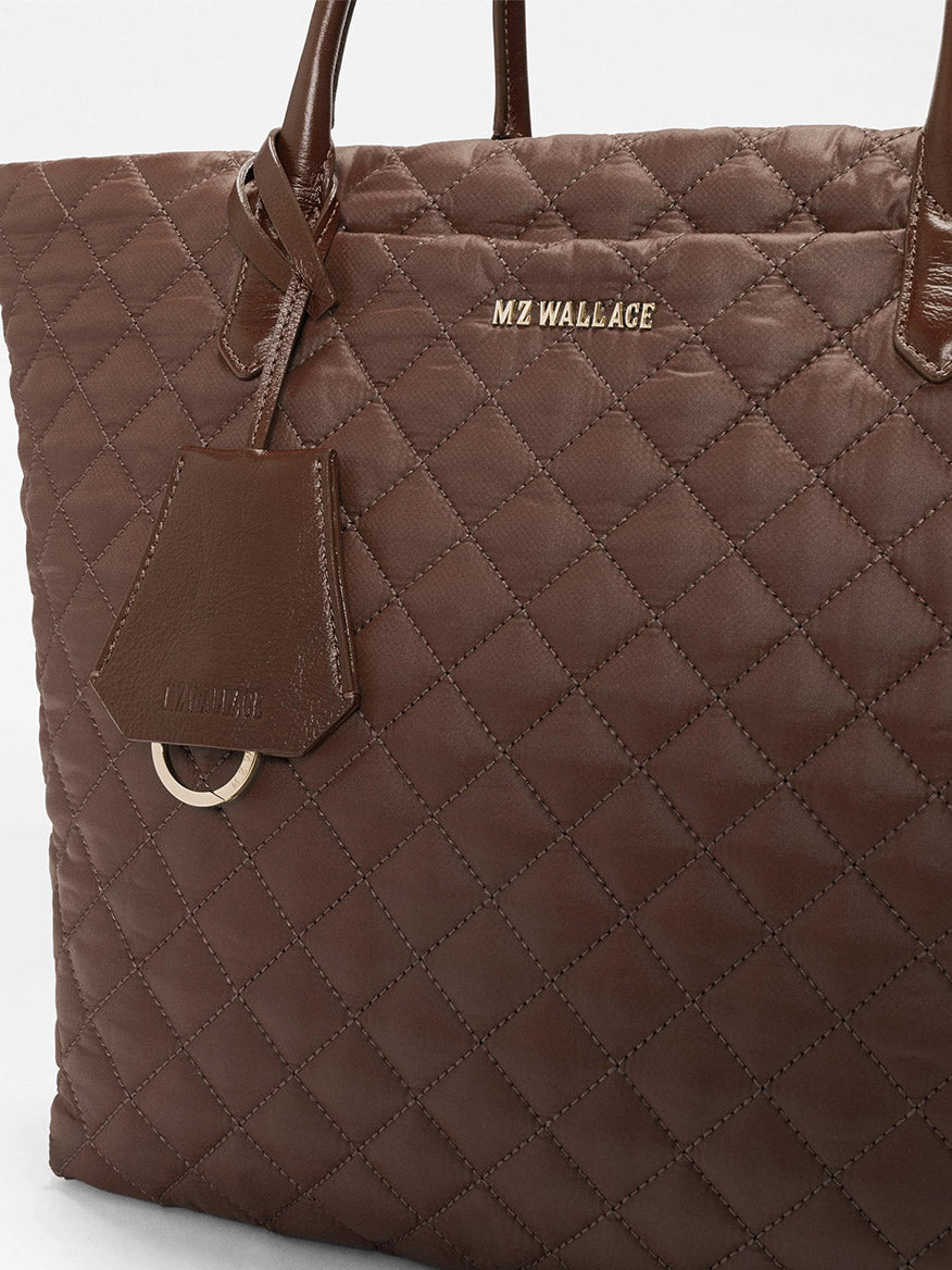 Close-up of the MZ Wallace Medium Daily Tote in Walnut Oxford, featuring brown quilted recycled nylon, “MZ WALLACE” gold lettering, and a matching leather tag attached to one handle.