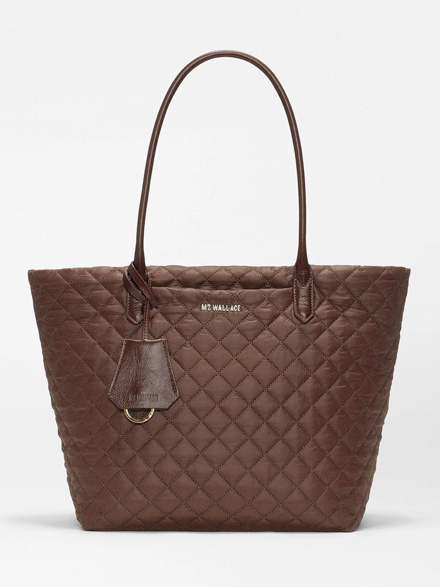 Introducing the MZ Wallace Medium Daily Tote in Walnut Oxford—a brown quilted bag made from recycled nylon, featuring two shoulder straps, a luggage tag, and gold-tone hardware. The brand name “MZ WALLACE” is displayed on the front. This spacious tote is perfect for carrying your laptop and daily essentials.