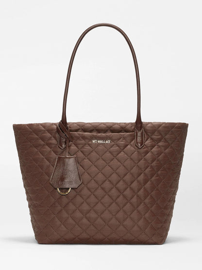 Introducing the MZ Wallace Medium Daily Tote in Walnut Oxford—a brown quilted bag made from recycled nylon, featuring two shoulder straps, a luggage tag, and gold-tone hardware. The brand name “MZ WALLACE” is displayed on the front. This spacious tote is perfect for carrying your laptop and daily essentials.