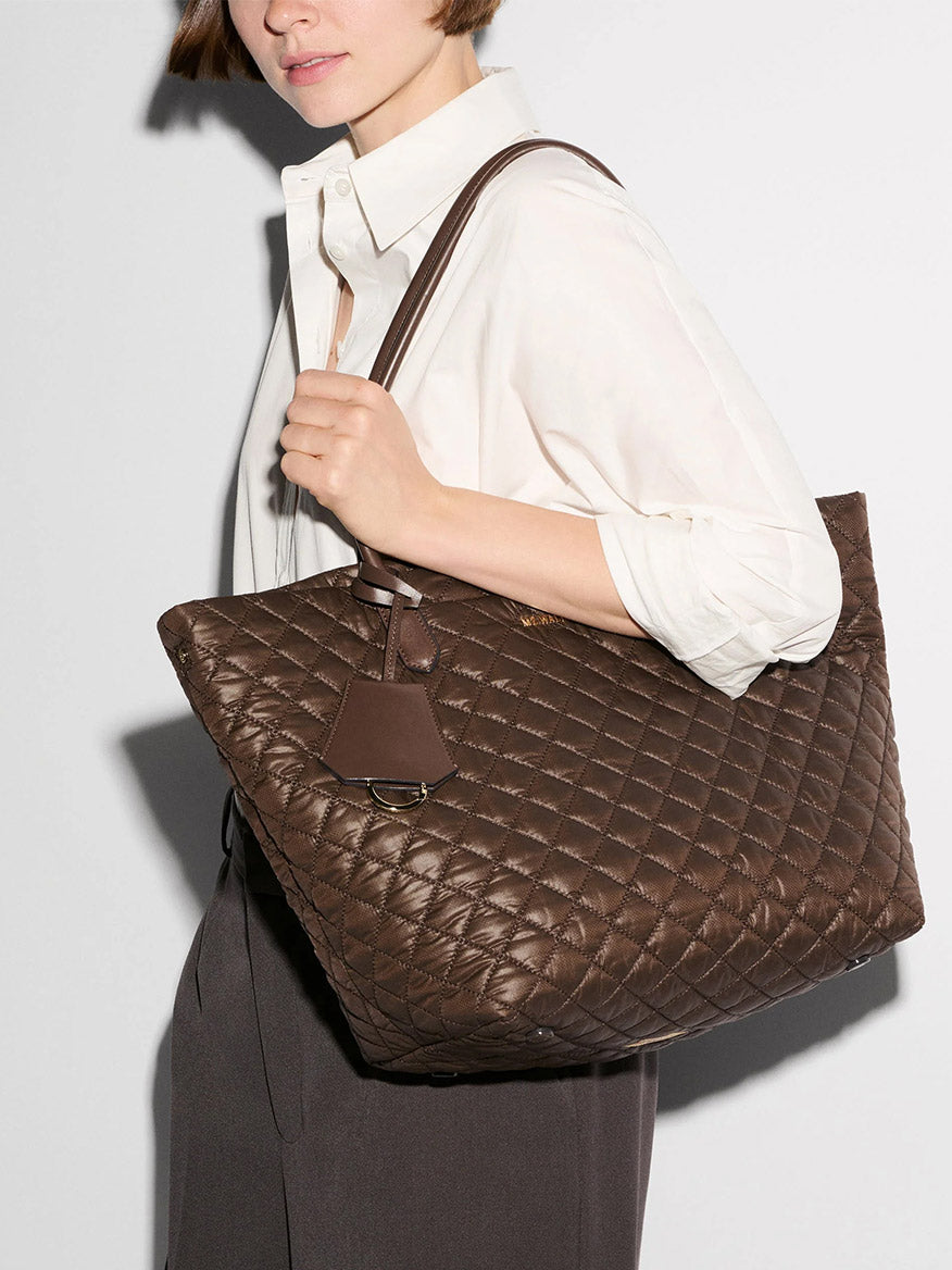 A woman wearing a white shirt and dark trousers holds the MZ Wallace Medium Daily Tote in Walnut Oxford over her shoulder against a plain background.