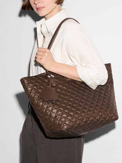 A woman wearing a white shirt and dark trousers holds the MZ Wallace Medium Daily Tote in Walnut Oxford over her shoulder against a plain background.