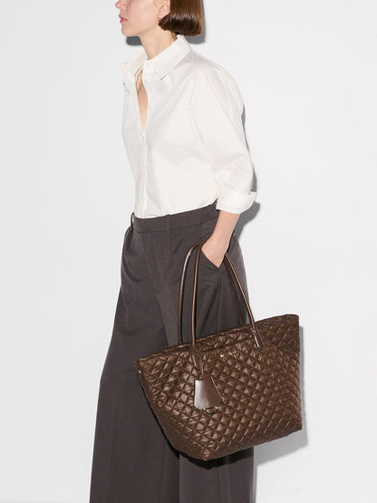 Woman wearing a white button-up shirt and dark wide-leg pants, holding the MZ Wallace Medium Daily Tote in Walnut Oxford—a spacious, quilted brown recycled nylon tote with two handles, a leather tag, and ample room to serve perfectly as a laptop tote bag.