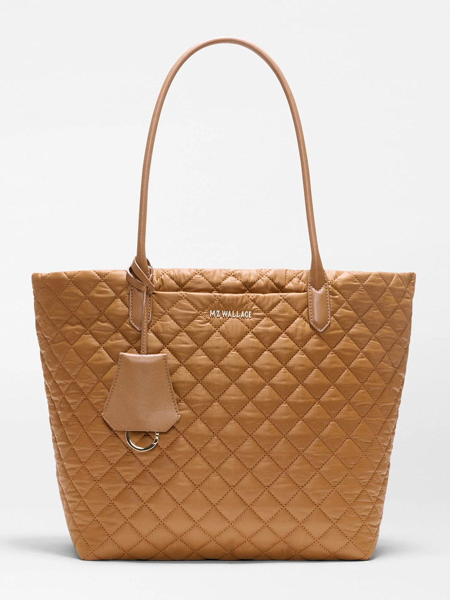 The MZ Wallace Medium Daily Tote in Cognac Oxford features a tan quilted design with two handles, a small hanging tag, and the “MZ WALLACE” logo on the front. Made from lightweight quilted nylon, this versatile carryall is displayed against a plain light background.