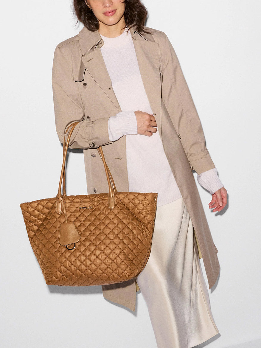 A woman wearing a beige trench coat and white outfit carries the MZ Wallace Medium Daily Tote in Cognac Oxford—a large, lightweight quilted nylon carryall.