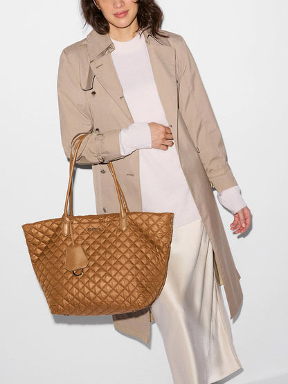 A woman wearing a beige trench coat and white outfit carries the MZ Wallace Medium Daily Tote in Cognac Oxford—a large, lightweight quilted nylon carryall.