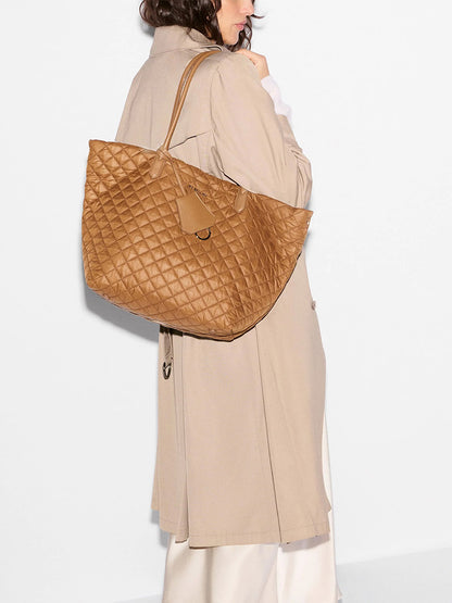 A woman in a beige trench coat carries the MZ Wallace Medium Daily Tote in Cognac Oxford—a large, lightweight quilted nylon bag—over her shoulder against a plain white background, showcasing an effortlessly chic everyday carryall.