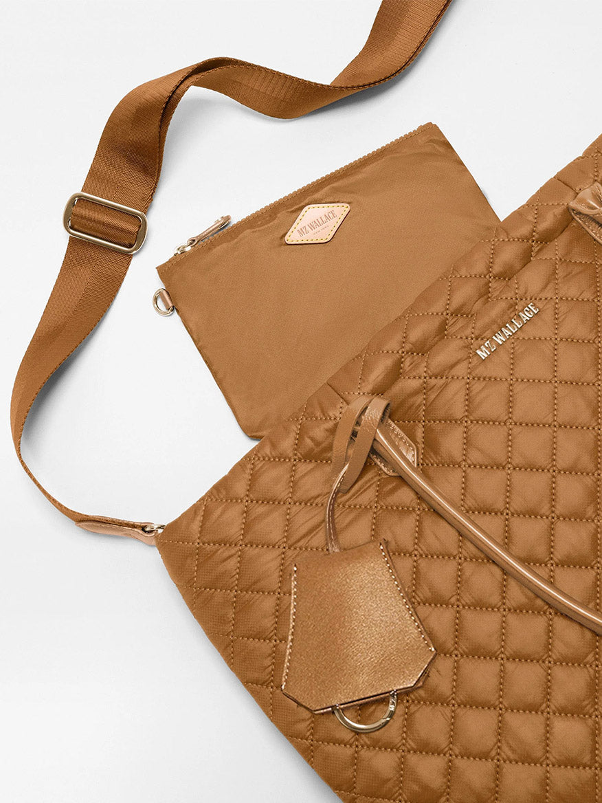 The MZ Wallace Medium Daily Tote in Cognac Oxford is a lightweight quilted nylon shoulder bag in tan, featuring an adjustable strap, matching pouch, and metallic tag—all presented on a white background. Inspired by the classic cognac tote, it’s the ideal everyday carryall.