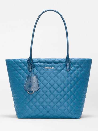 The MZ Wallace Medium Daily Tote in Atlantic Blue Oxford features a blue quilted recycled nylon exterior, two handles, a small attached leather tag, and the “MZ WALLACE” logo on the front. Set against a plain white background, it’s an ideal choice for your everyday Metro Daily Tote or as a laptop tote.
