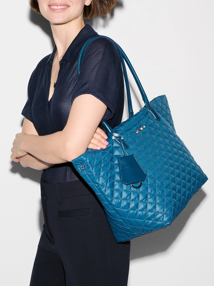 A woman in a navy shirt holds the MZ Wallace Medium Daily Tote in Atlantic Blue Oxford, featuring top handles, a tag, and a dedicated laptop sleeve, standing against a plain light background.