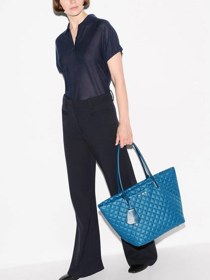 A person wearing a navy blouse and navy pants stands holding the MZ Wallace Medium Daily Tote in Atlantic Blue Oxford, which features top handles, is crafted with recycled nylon, and includes an interior laptop sleeve.