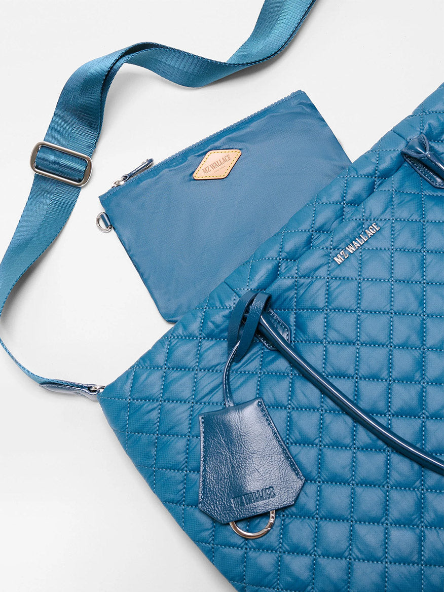MZ Wallace Medium Daily Tote in Atlantic Blue Oxford crafted from recycled nylon, showcasing a quilted design with a matching blue strap and leather tag. Pictured with a small blue zippered pouch featuring a beige emblem, this stylish tote includes a convenient laptop sleeve for your daily essentials.
