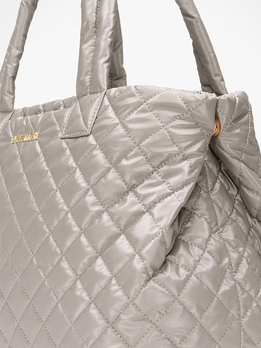 Close-up of the MZ Wallace Medium East West Metro Tote in Sable Pearl Metallic Oxford, featuring quilted silver-gray fabric, padded handles, Italian leather trim, a gold logo on the top left, and an optional crossbody strap for versatility.