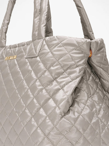 Close-up of the MZ Wallace Medium East West Metro Tote in Sable Pearl Metallic Oxford, featuring quilted silver-gray fabric, padded handles, Italian leather trim, a gold logo on the top left, and an optional crossbody strap for versatility.