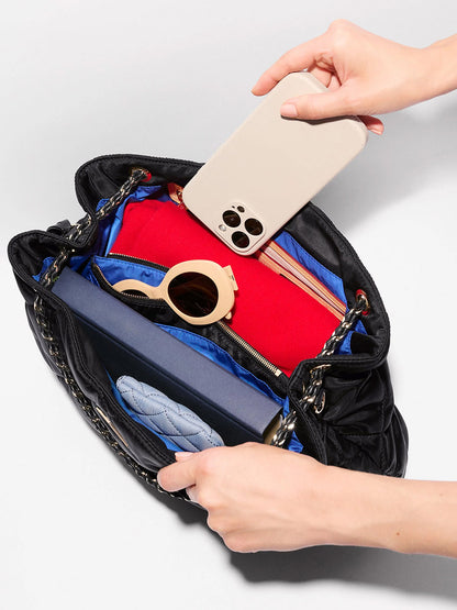 A person places a smartphone into an open MZ Wallace Medium Frankie Tote in Black Bedford, a quilted nylon handbag containing sunglasses, a blue wallet, and a closed book, featuring an adjustable crossbody strap.