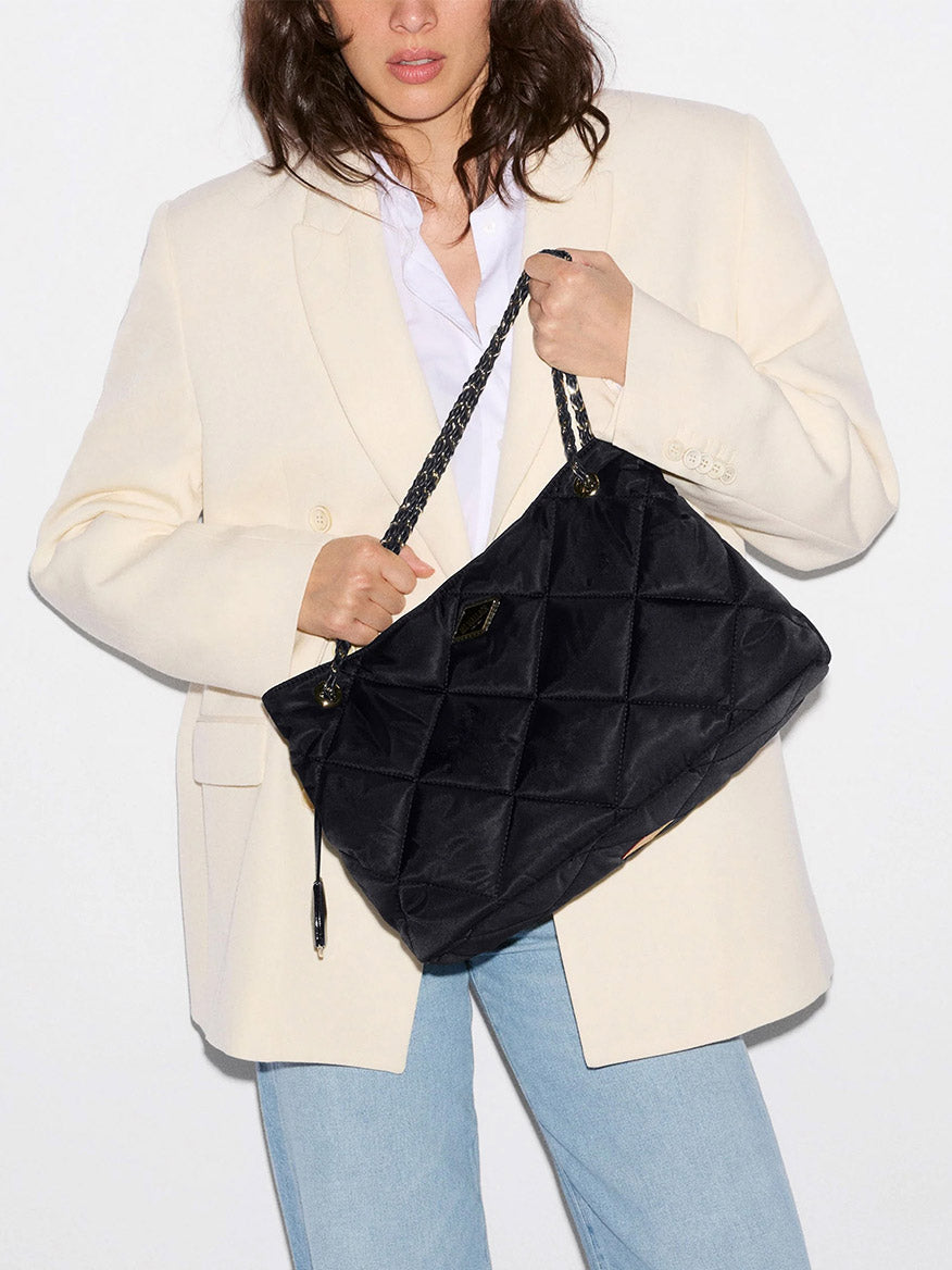 Woman in a cream blazer and blue jeans holding an MZ Wallace Medium Frankie Tote in Black Bedford—a large quilted nylon bag with chain straps and an adjustable crossbody strap—against a plain background.
