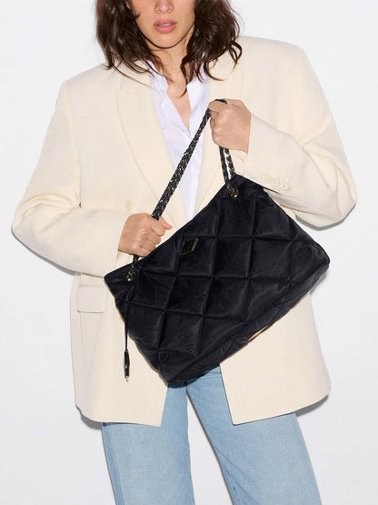 Woman in a cream blazer and blue jeans holding an MZ Wallace Medium Frankie Tote in Black Bedford—a large quilted nylon bag with chain straps and an adjustable crossbody strap—against a plain background.