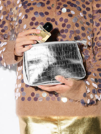 Person holding the MZ Wallace Medium Luna Cosmetic in Silver Croc Effect and a perfume bottle, wearing a fuzzy sequin top and gold metallic pants.