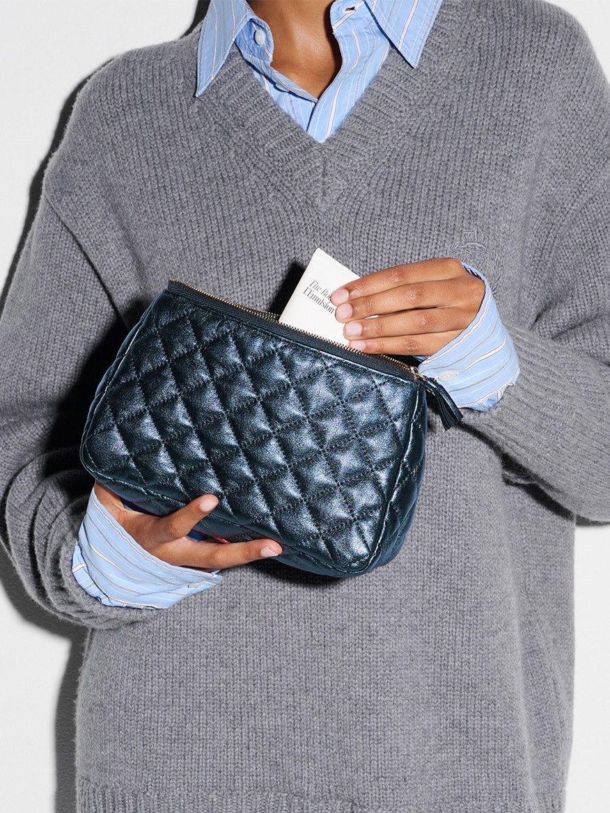 A person in a grey sweater and blue striped shirt holds the MZ Wallace Medium Luna Cosmetic in Midnight Metallic Leather and inserts a white card into it.