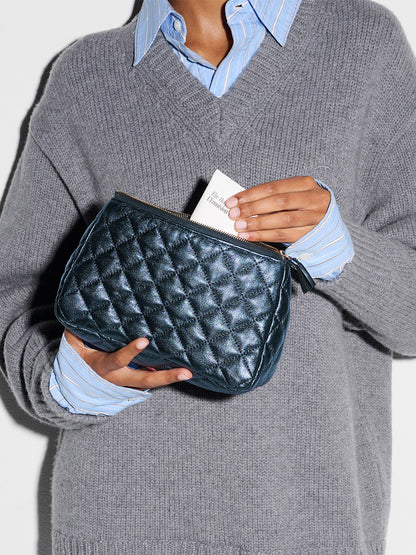 A person in a grey sweater and blue striped shirt holds the MZ Wallace Medium Luna Cosmetic in Midnight Metallic Leather and inserts a white card into it.
