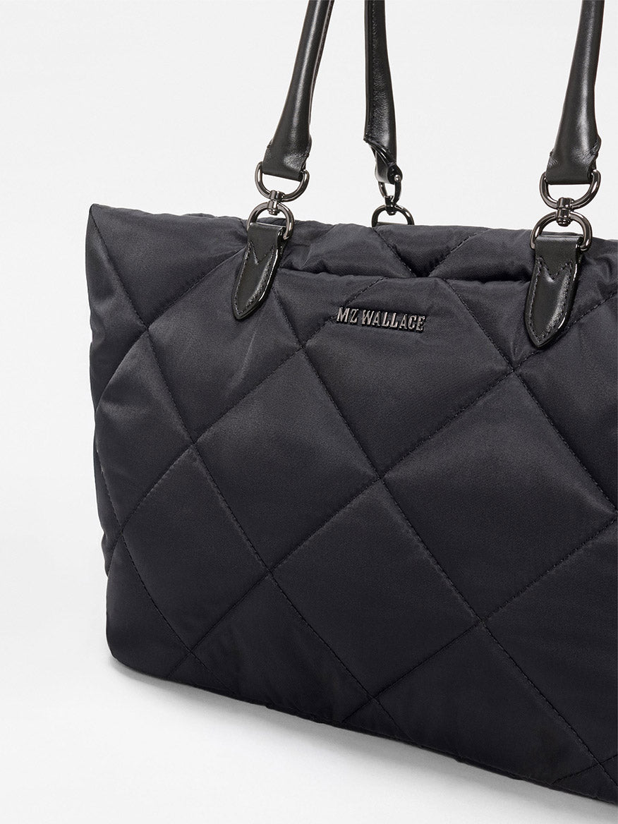 A close-up of the MZ Wallace Medium Madison E-W Tote in Black Bedford, featuring quilted design, Italian leather trim, black leather handles, and silver hardware against a plain background.