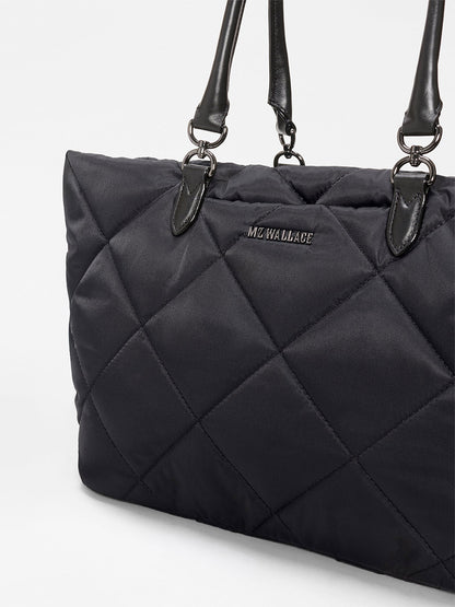 A close-up of the MZ Wallace Medium Madison E-W Tote in Black Bedford, featuring quilted design, Italian leather trim, black leather handles, and silver hardware against a plain background.