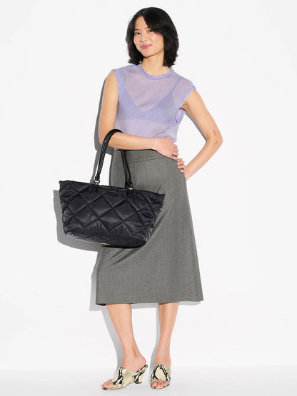 A woman stands against a white background wearing a sheer lavender top, grey midi skirt, patterned heeled mules, and holding the MZ Wallace Medium Madison E-W Tote in Black Bedford with Italian leather trim.